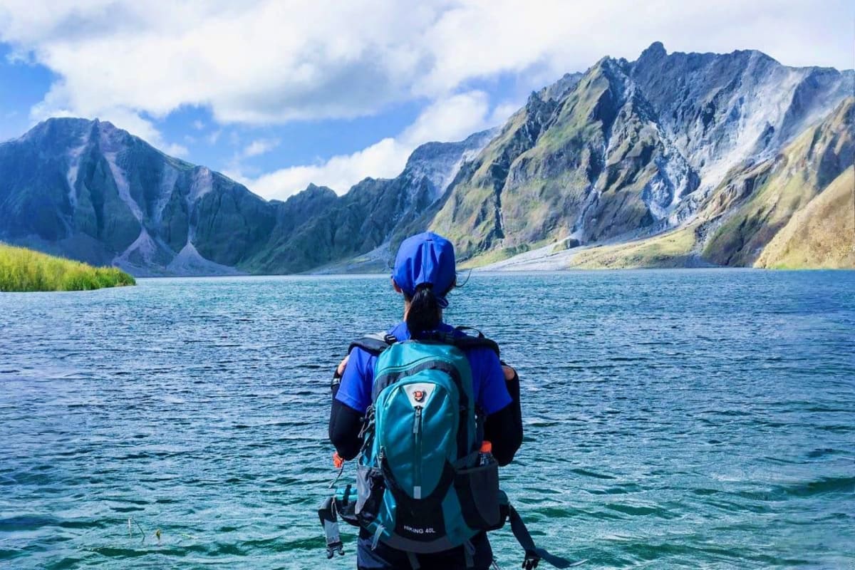 Manila Angeles Budget Tour Hiker wearing blue helmet and backpack standing in turquoise lake facing dramatic mountain peaks with rocky cliffs