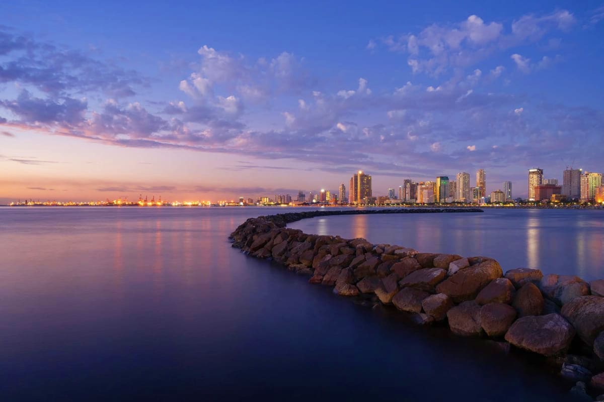 Exclusively Manila San Diego skyline at sunset with illuminated downtown buildings reflected in calm bay waters and rocky jetty in foreground