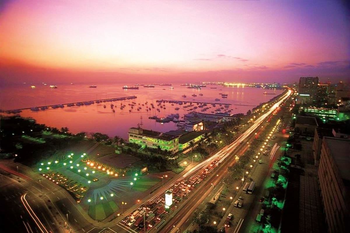 Exclusively Manila Aerial sunset view of Manila Bay with illuminated waterfront promenade, parking lot, and anchored vessels at dusk