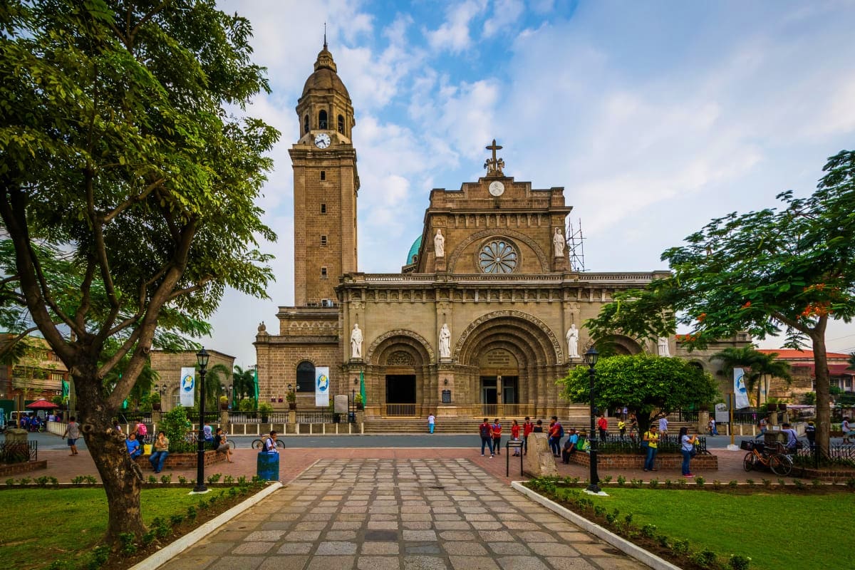 Exclusively Manila Historic stone cathedral with tall bell tower in Manila plaza, surrounded by trees and visitors
