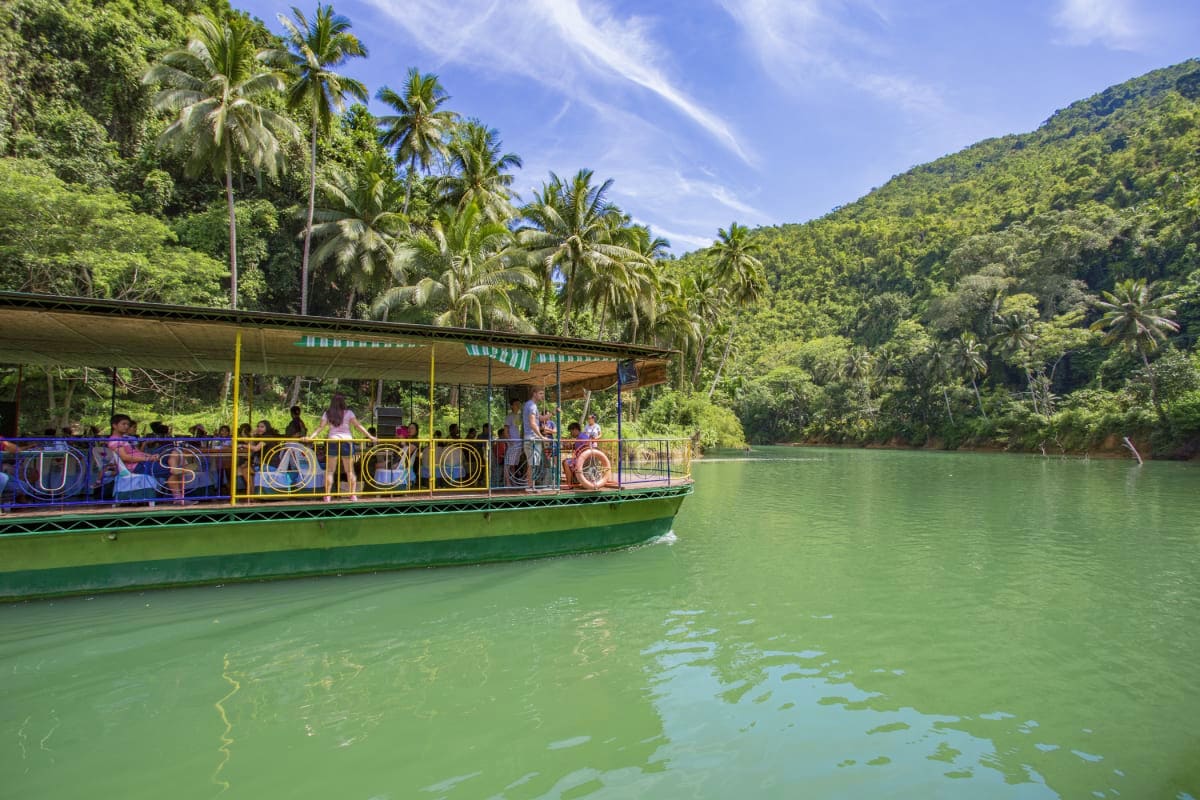 Simply Bohol Tourist boat with passengers on turquoise river surrounded by palm trees and forested hills in Bohol, Philippines