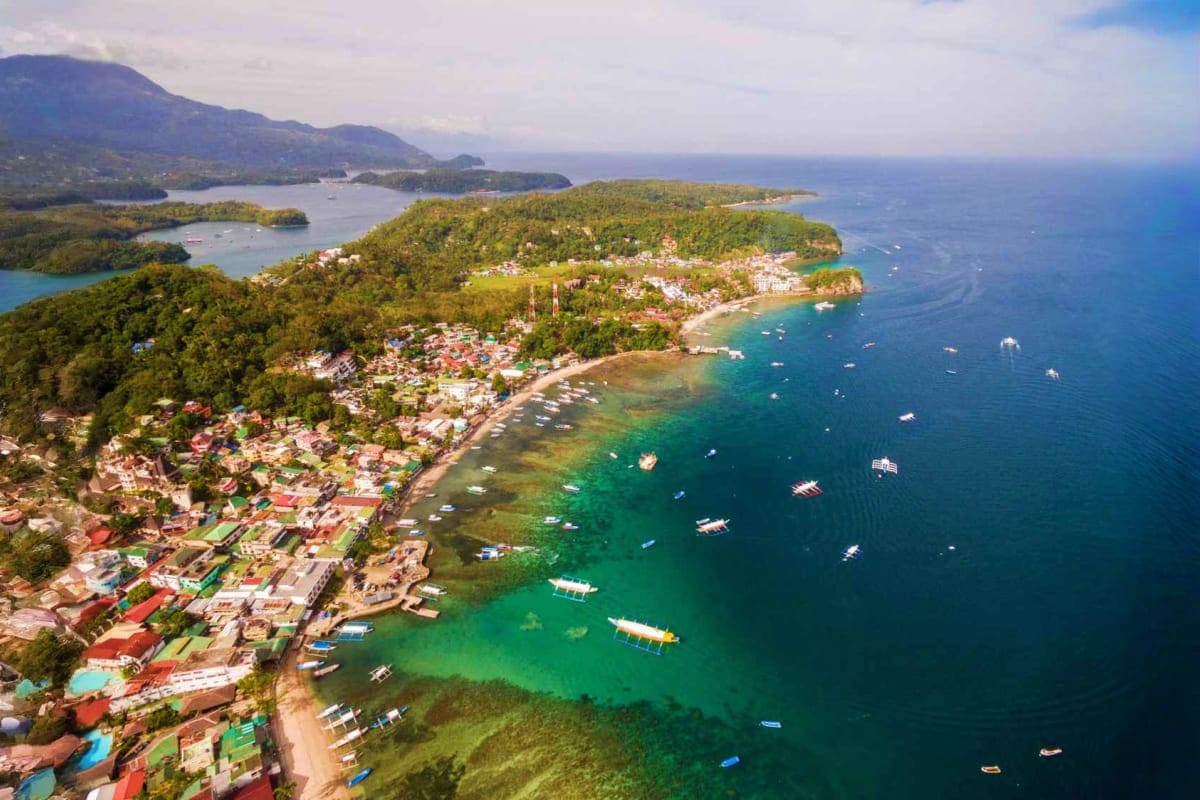 Puerto Galera Diving Tour Aerial view of Puerto Galera coastal town with turquoise waters, anchored boats, and forested hillside surrounded by mountain