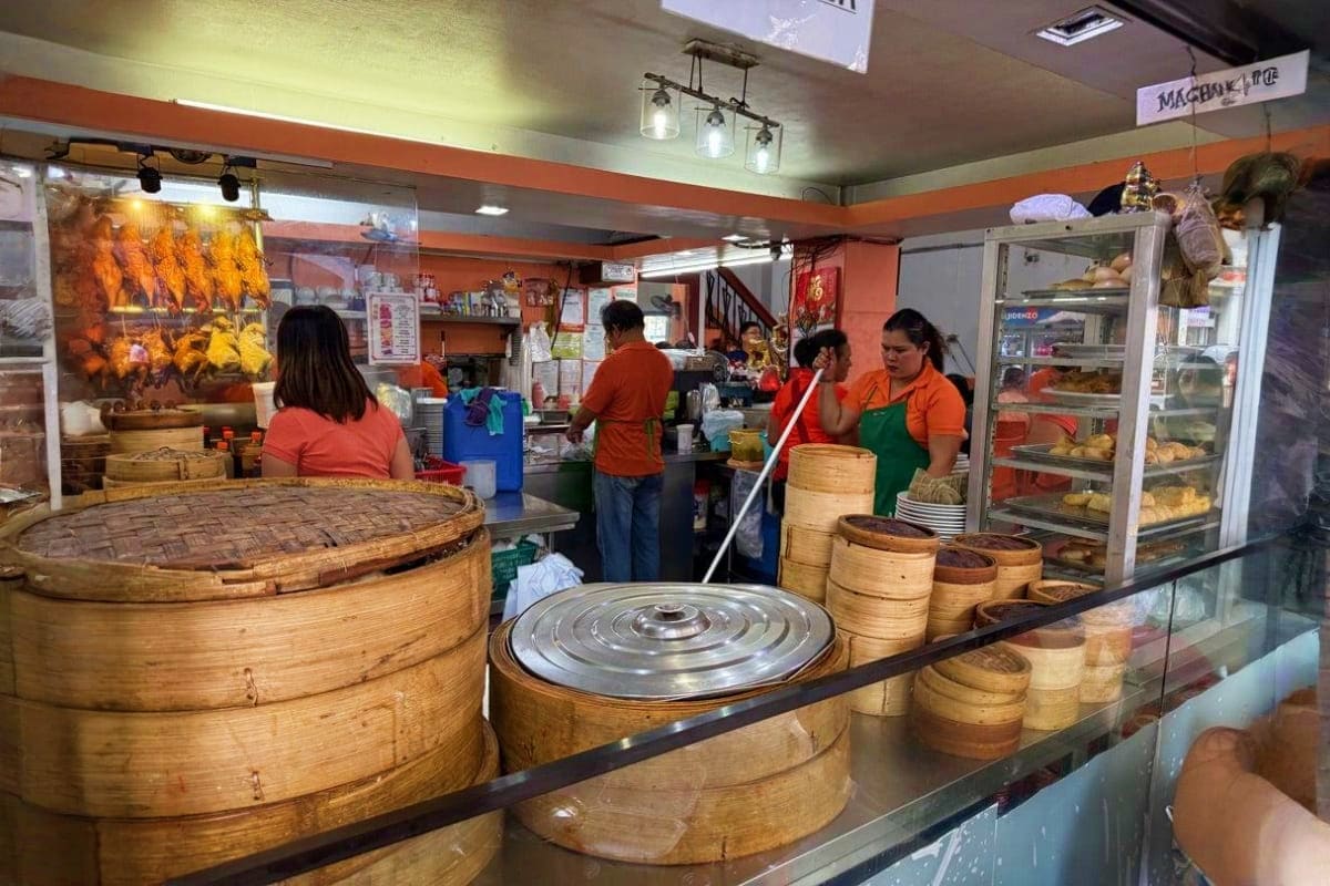 Manila Food Tour Busy Asian dim sum restaurant kitchen with bamboo steamers, hanging roasted ducks, staff in orange uniforms, and steamed bask