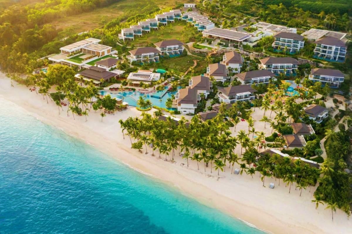 Island Escape Package Aerial view of beachfront resort with white sand beach, turquoise ocean, palm trees, villas, and blue swimming pool