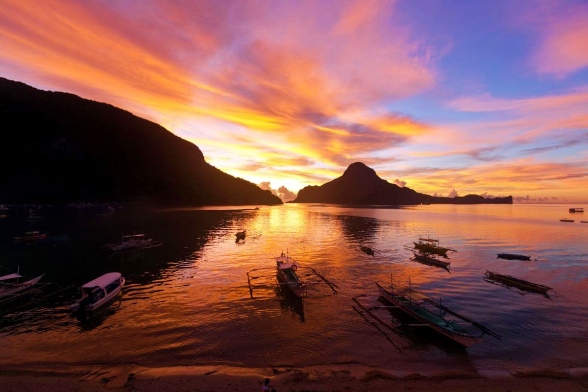 El Nido Escape Tour Tropical beach at sunset with traditional boats anchored in calm waters, limestone cliffs silhouetted against golden sky in E