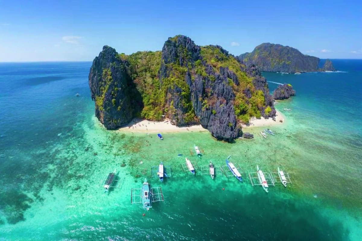 El Nido Escape Tour Aerial view of limestone cliffs with white sand beach, turquoise lagoon, and traditional boats anchored in crystal-clear wate