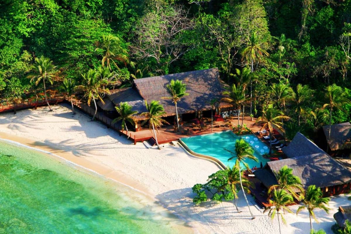 El Nido Escape Tour Aerial view of beachfront resort with thatched-roof bungalows, turquoise pool, sandy beach, and dense tropical forest in El N