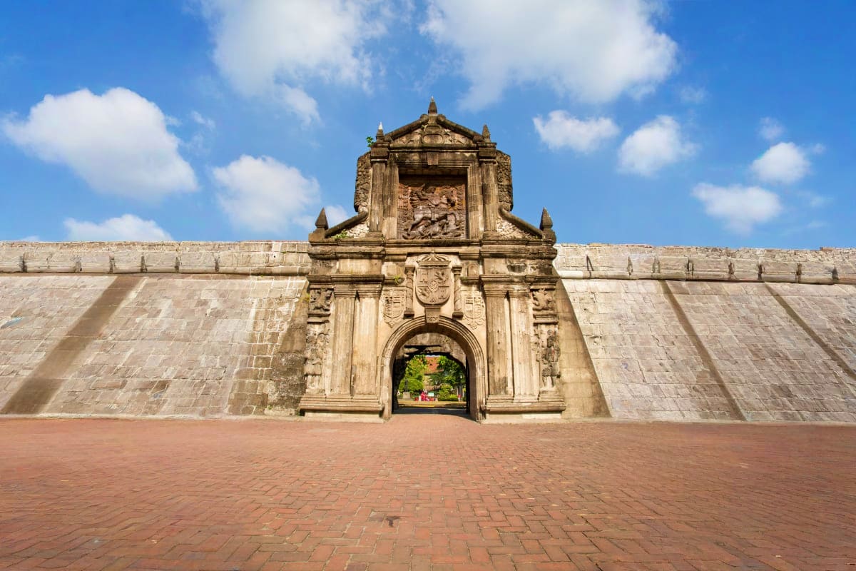Manila Coron Escape Tour Historic stone fortified gate with ornate carved archway and red brick courtyard under blue sky with clouds