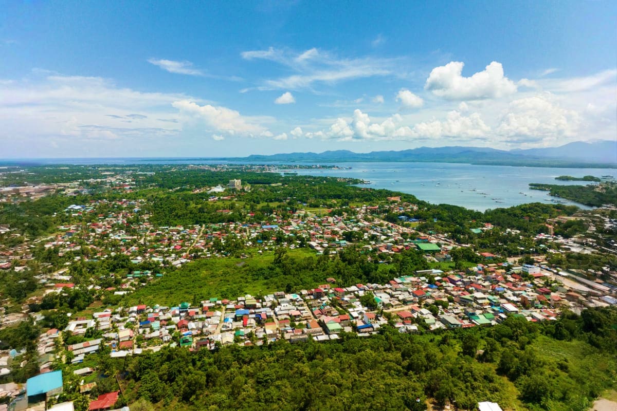Pristine Palawan Tour Aerial view of a coastal town nestled between lush green mountains and turquoise bay waters under a cloudy blue sky