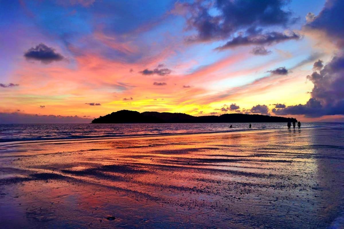Tropical beach sunset with silhouetted island in distance, shallow water reflecting golden and orange sky, people wading in s