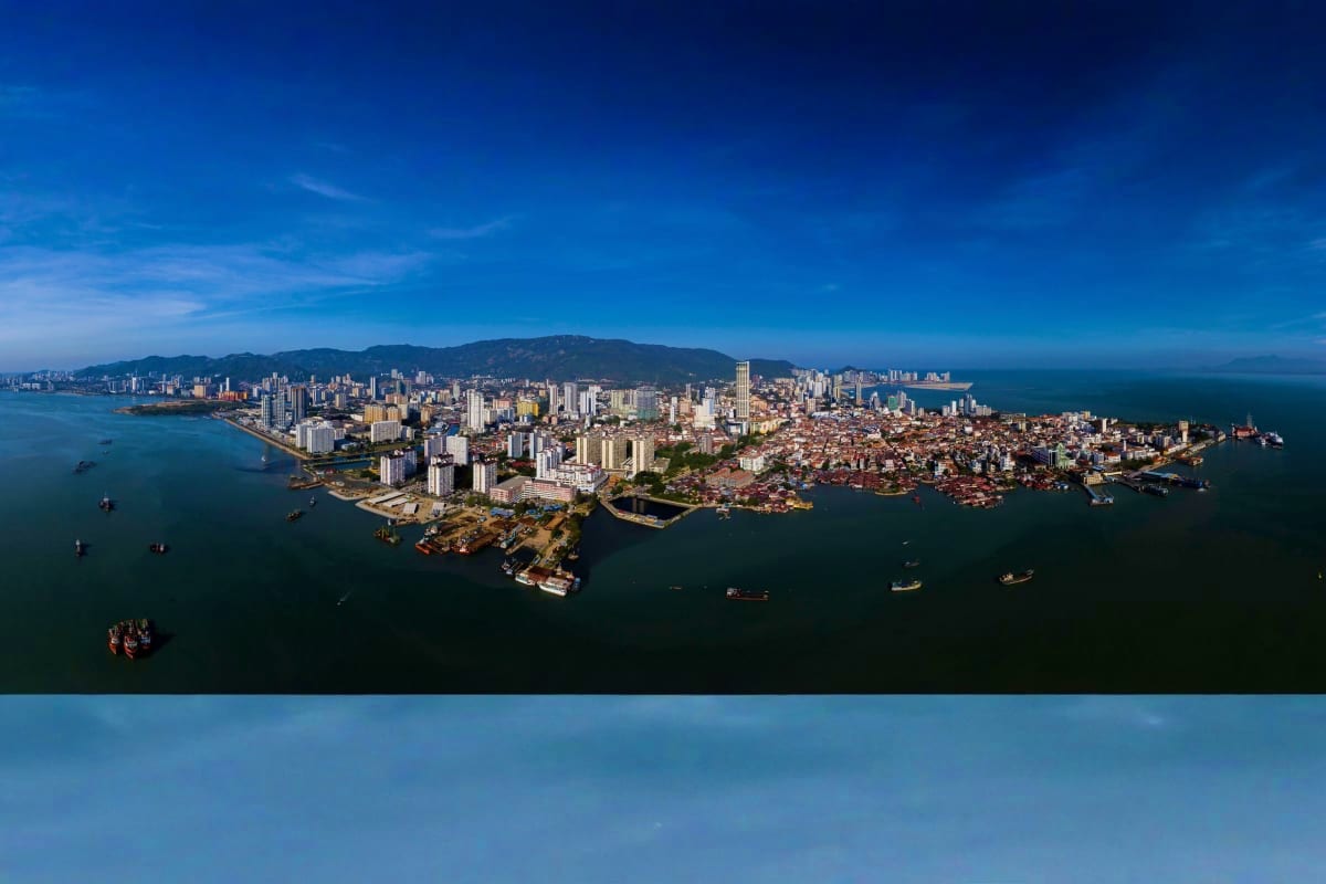 Aerial view of Penang cityscape with high-rise buildings, coastal neighborhoods, and turquoise waters surrounded by mountains