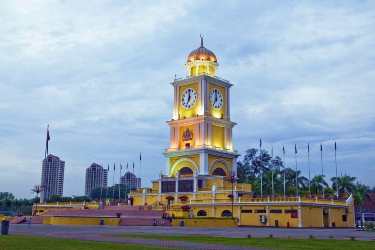 Illuminated yellow clock tower with golden dome at dusk, surrounded by flags and palm trees in Malaysia