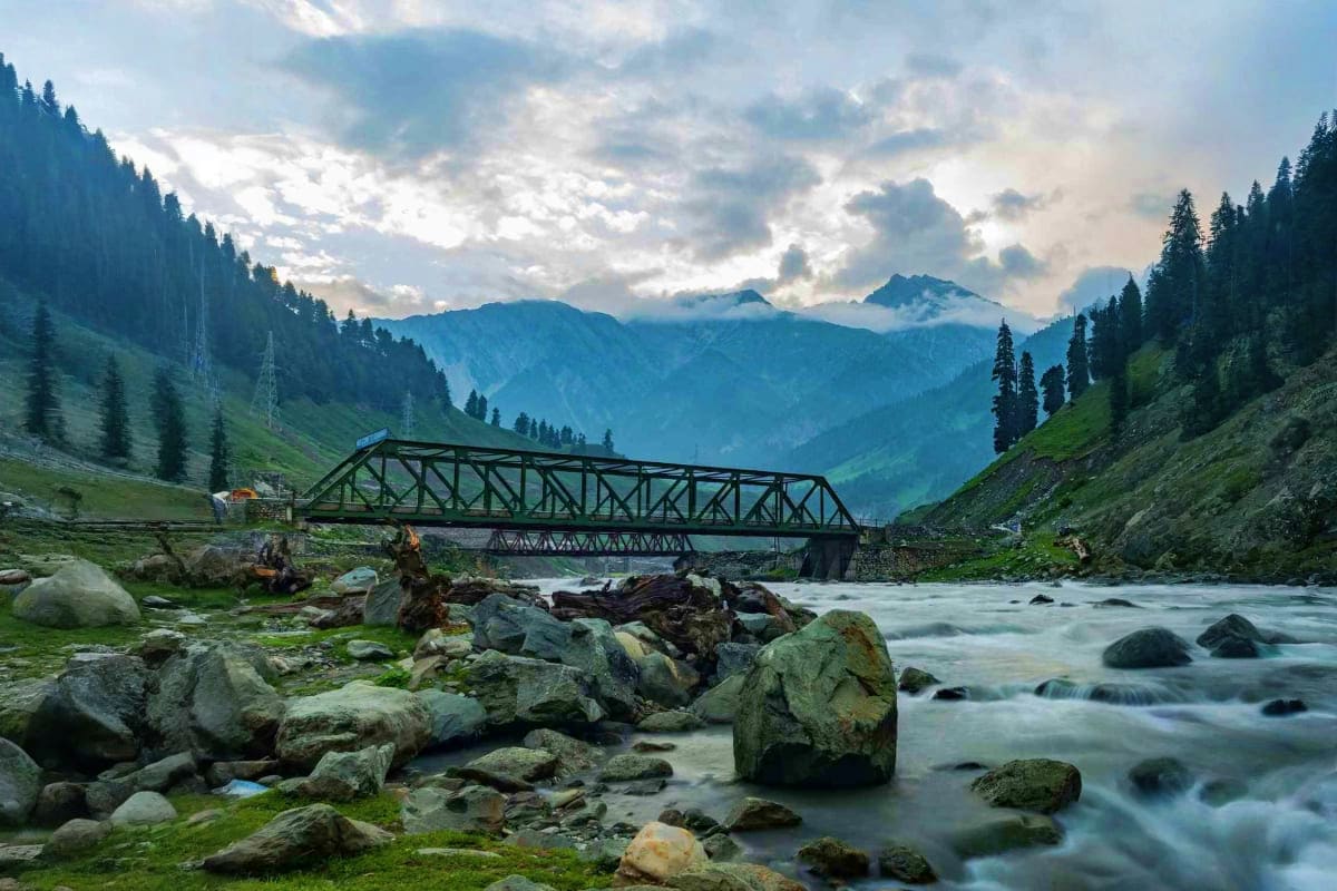 Steel truss bridge spanning mountain river in Kashmir valley with snow-capped peaks and evergreen forests in background