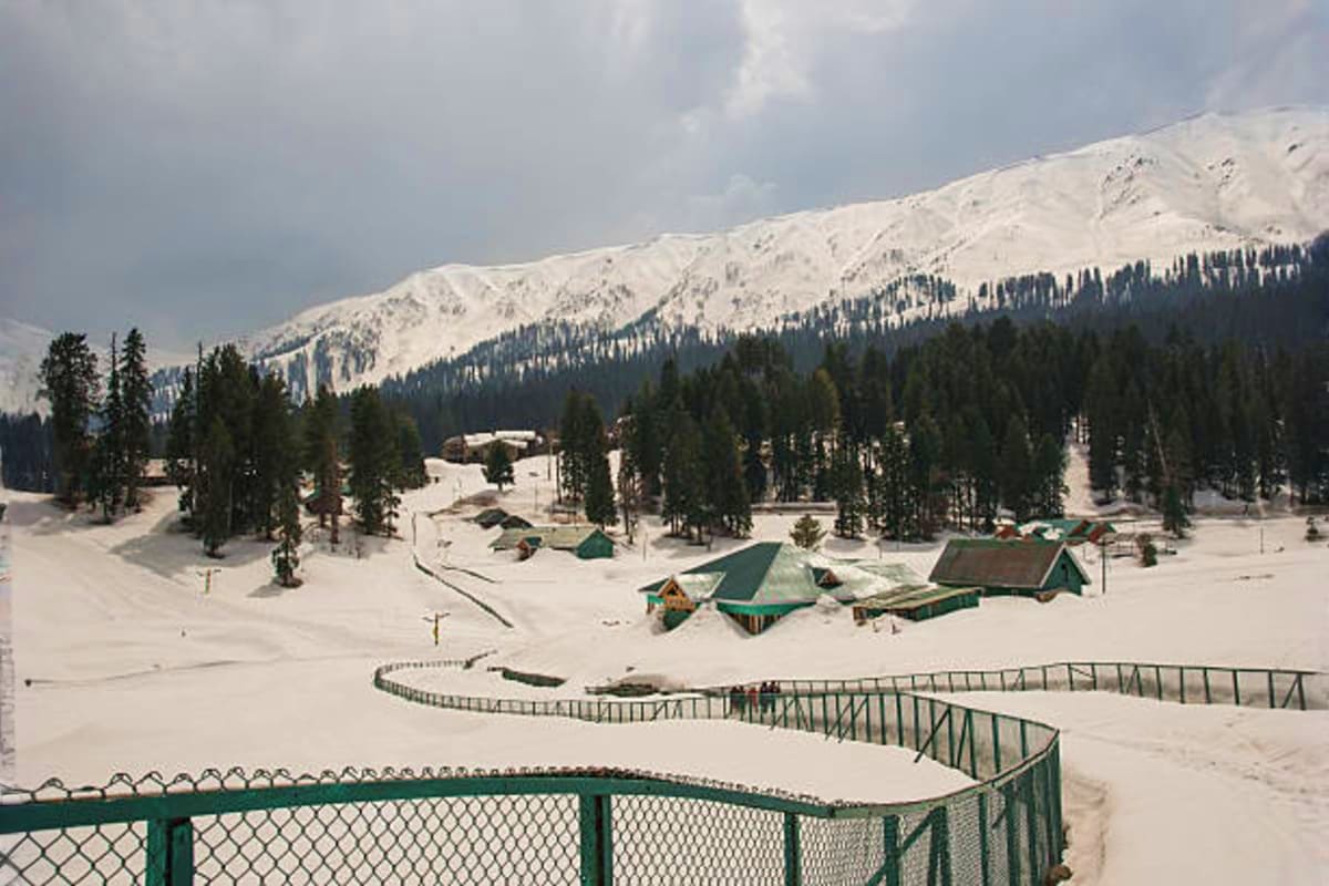 Snow-covered valley in Kashmir with pine forests, snow-capped mountains, green-roofed buildings, and metal fencing in foregro