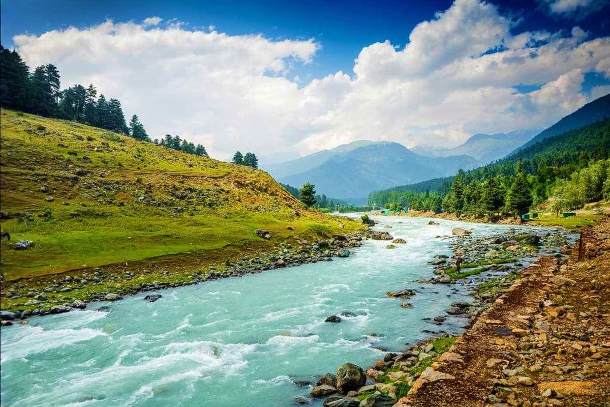 Turquoise mountain river flowing through lush green valley with forested slopes and snow-capped peaks under blue sky with whi