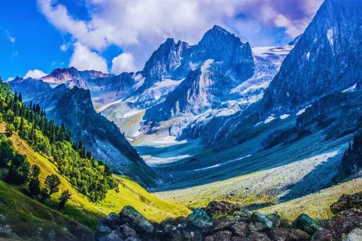 Alpine valley with snow-capped mountain peaks, green meadows, and rocky terrain under blue sky with clouds