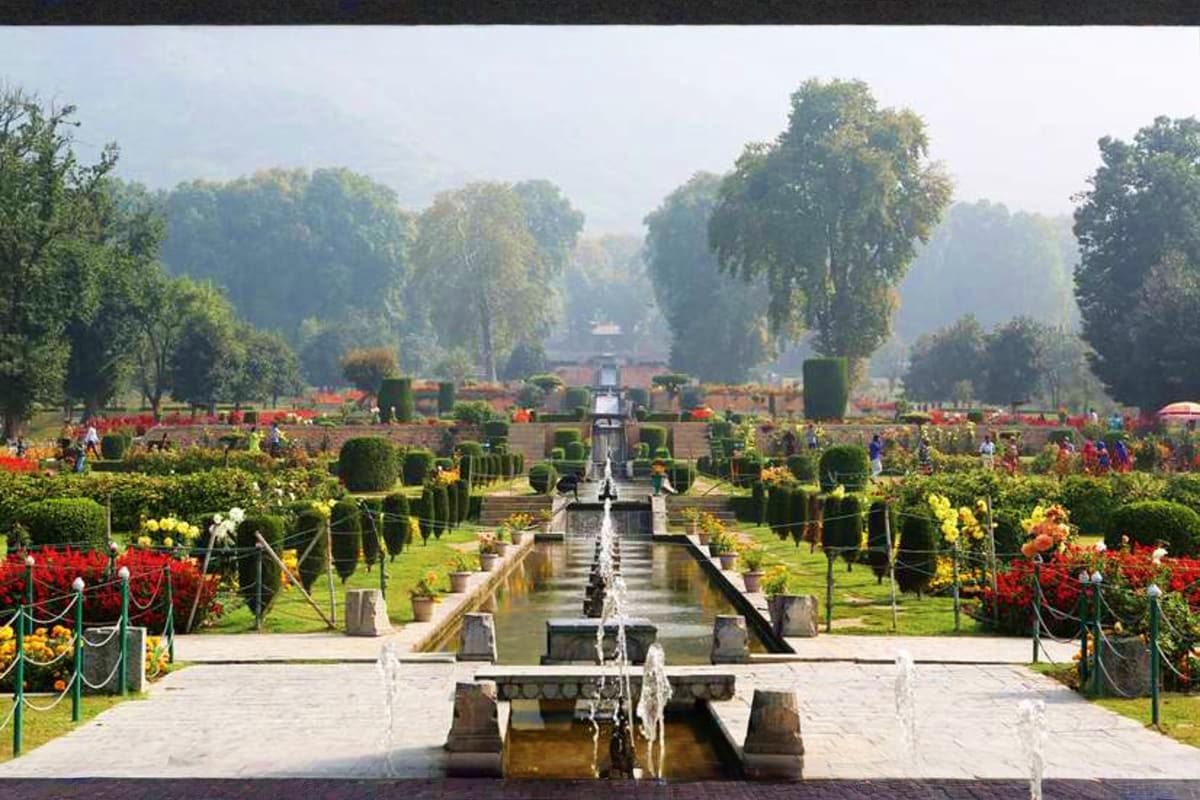 Formal Mughal garden with central water channel, fountains, manicured hedges, red and yellow flowers, and tree-lined pathways