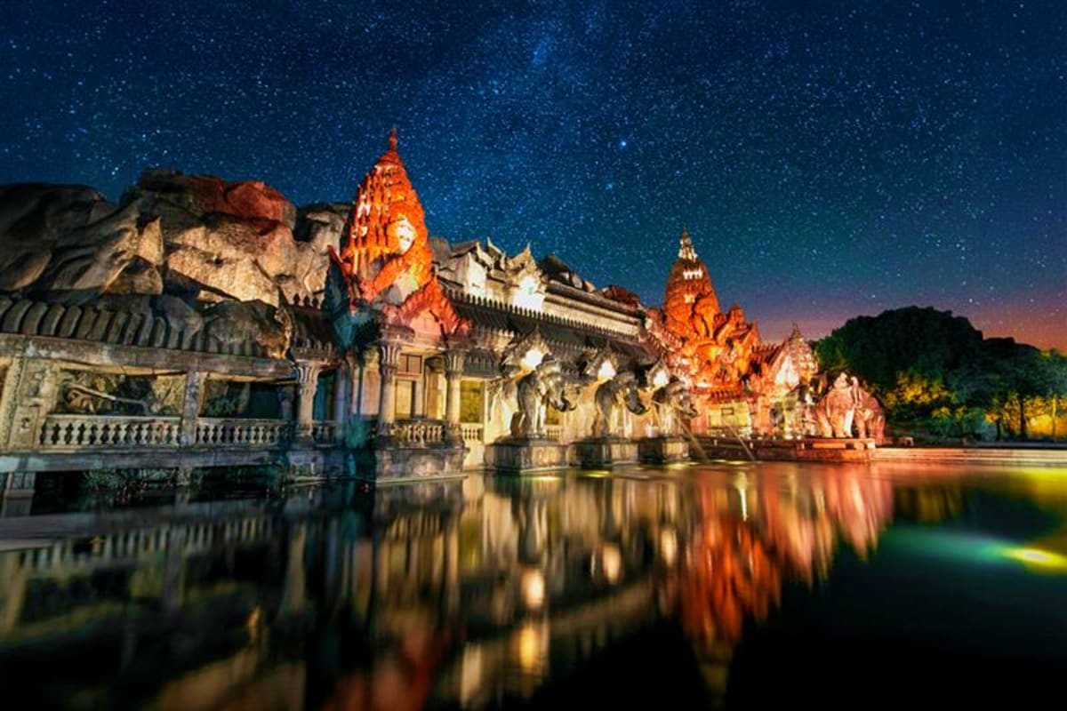 Illuminated Thai Buddhist temple with golden spires reflected in water at night under starry sky