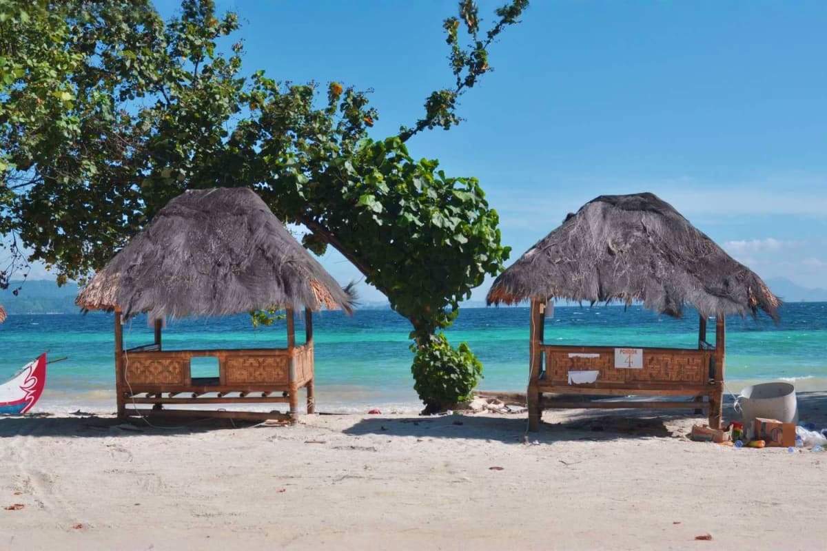 Bali Thrill & Serenity Two thatched-roof beach huts on white sand with turquoise ocean and tree in Bali, Indonesia