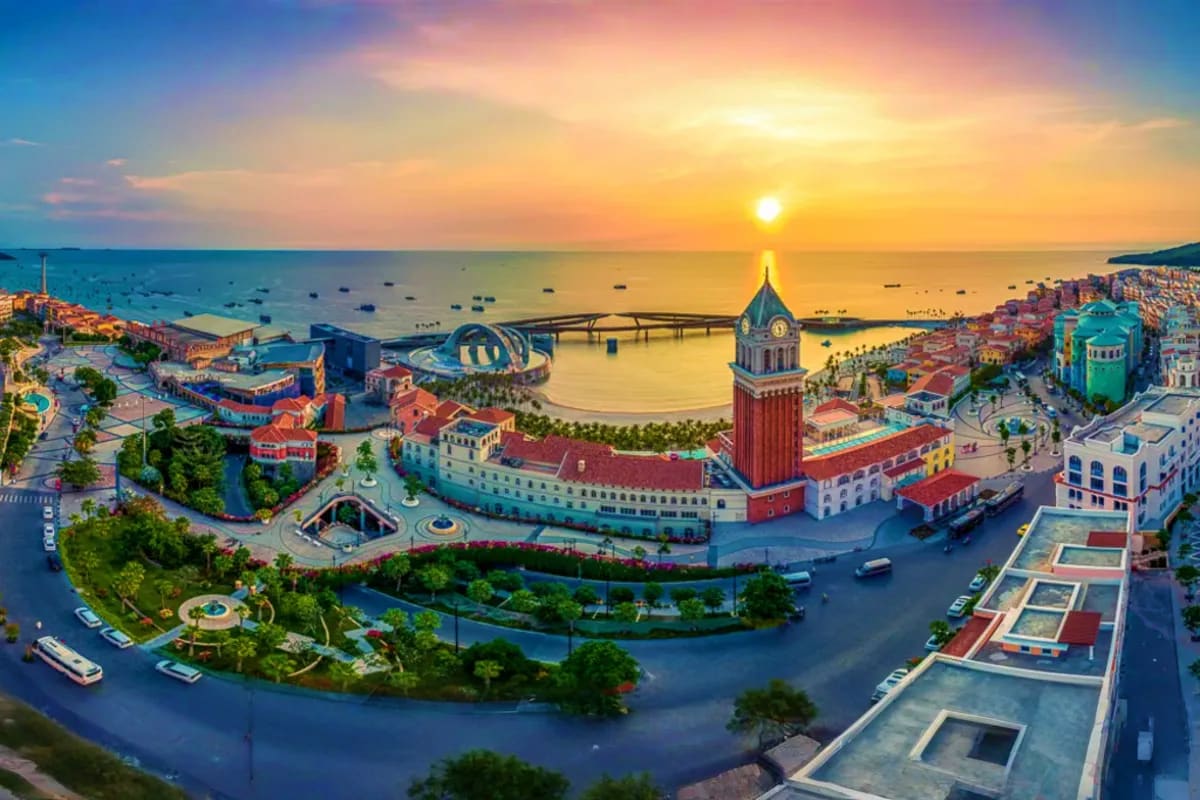 Aerial view of Phu Quoc Island resort at sunset with colorful buildings, bell tower, curved bridge, boats in harbor, and gold