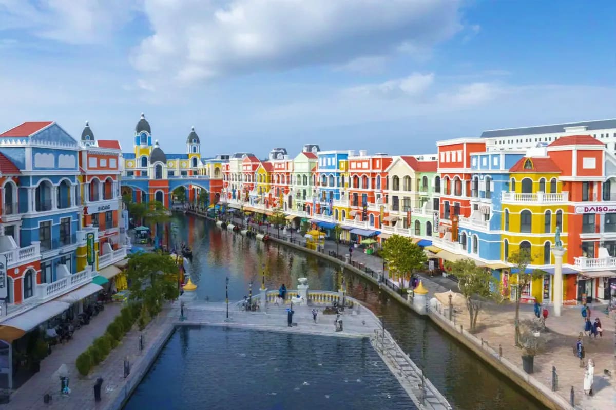 Colorful colonial-style buildings lining waterfront canals with pedestrians strolling along promenades under blue sky