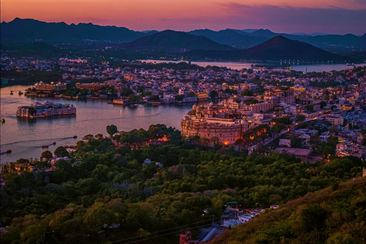 Aerial sunset view of Udaipur cityscape with illuminated palace on Lake Pichola, surrounded by mountains and residential buil