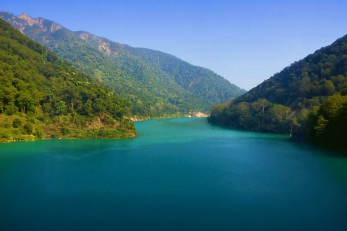 Turquoise alpine lake surrounded by dense green forested mountains under clear blue sky, scenic landscape destination
