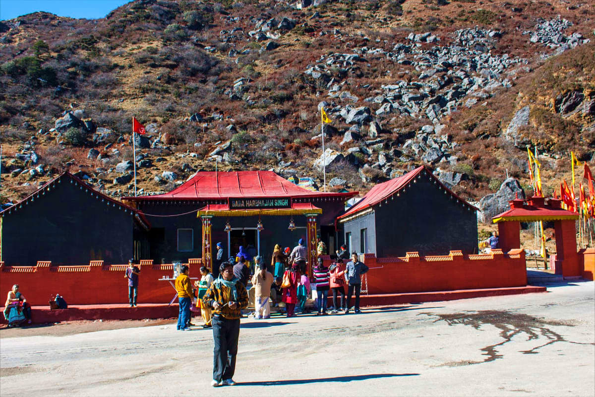 Visitors gathered at Naga Harhajangh Singh temple with red and black architecture nestled against rocky hillside in Himalayan
