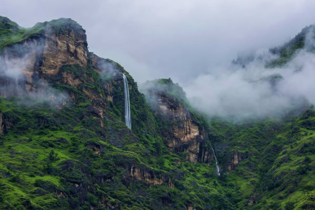 Misty mountain valley with waterfall cascading down rocky cliff face surrounded by lush green vegetation and low-lying clouds