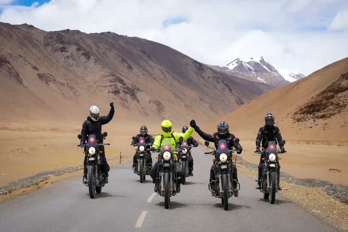 Group of five motorcycle riders in helmets and black gear riding on a mountain road with snow-capped peaks and barren valleys