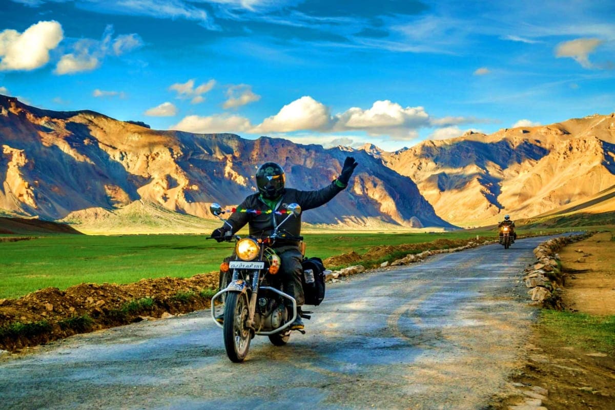 Motorcyclist with raised arm riding on mountain road in Ladakh valley with golden brown peaks and green fields under blue sky