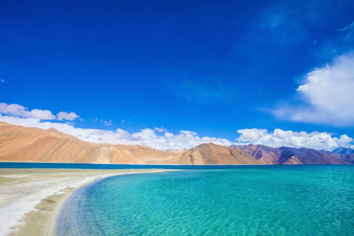 Turquoise waters of Pangong Lake with white sandy beach and barren brown mountains under blue sky with white clouds, Ladakh