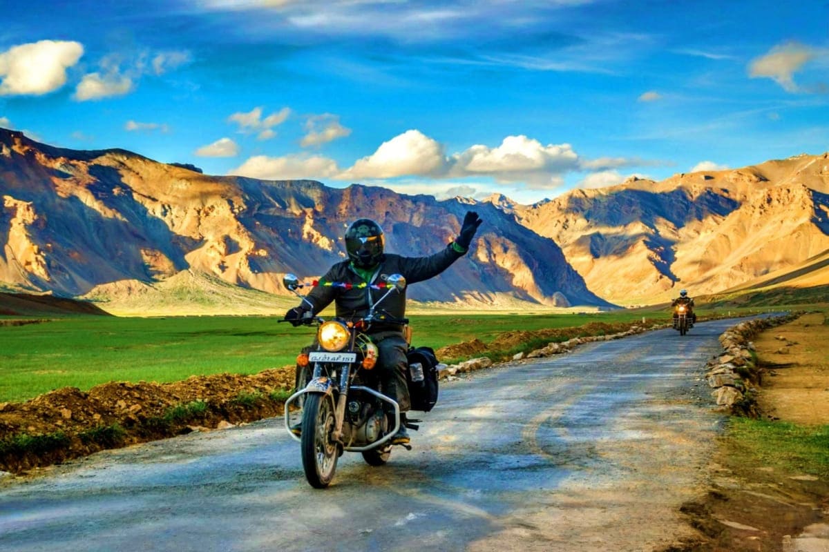Motorcyclist with raised arm riding on mountain road through Ladakh valley with dramatic brown peaks and green fields under b