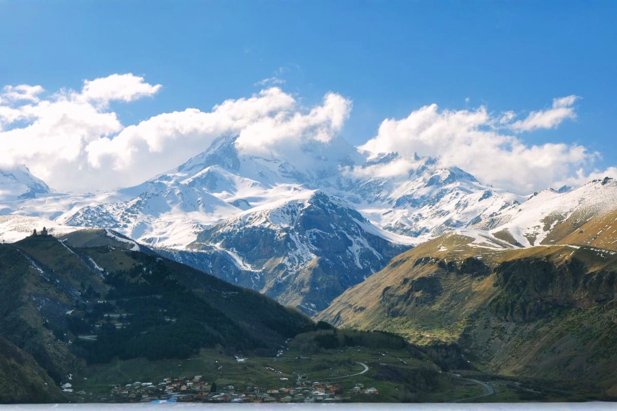 Snow-capped Himalayan mountains with green valley and village settlements below under blue sky with white clouds