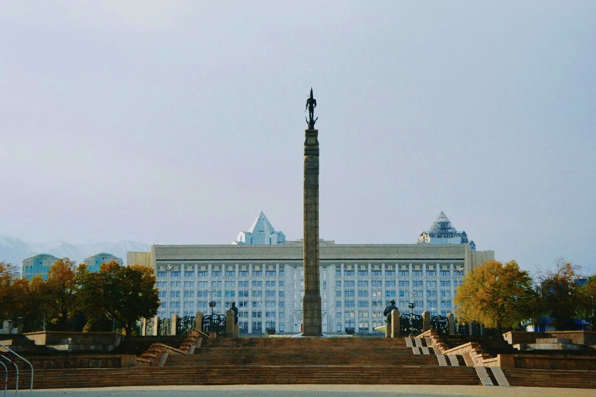 Tall monument with statue on top standing in front of large blue government building in central Asian city square with autumn