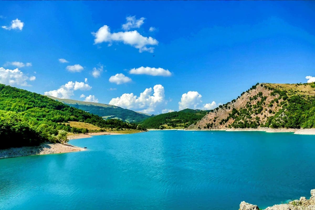 Turquoise alpine lake surrounded by forested mountains and rocky cliffs under blue sky with white clouds