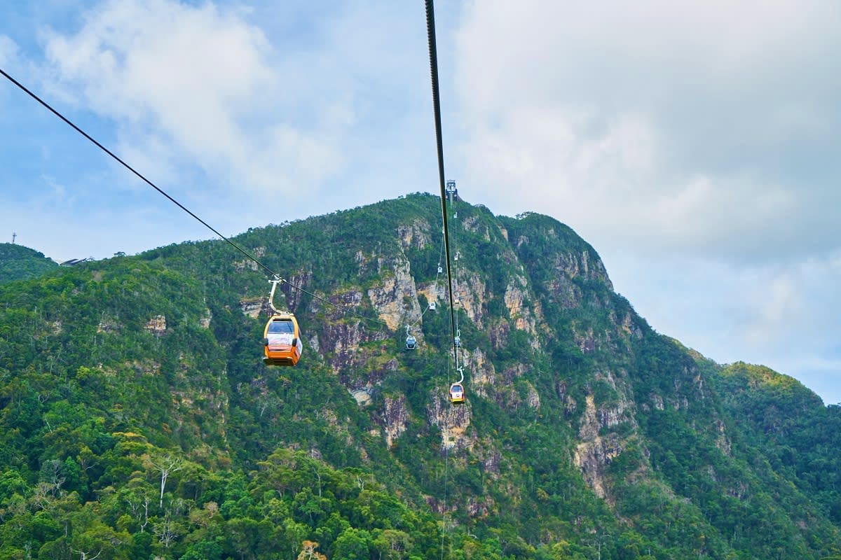 Cable car gondolas suspended over forested limestone mountains with lush green vegetation and rocky cliffs.