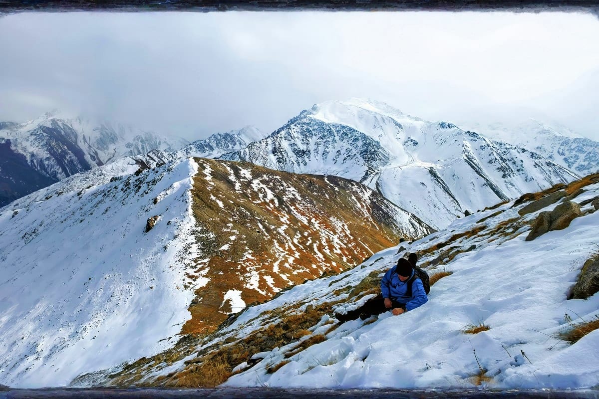 Hiker in blue jacket on snowy mountainside with snow-capped peaks in background, Kazakhstan alpine landscape