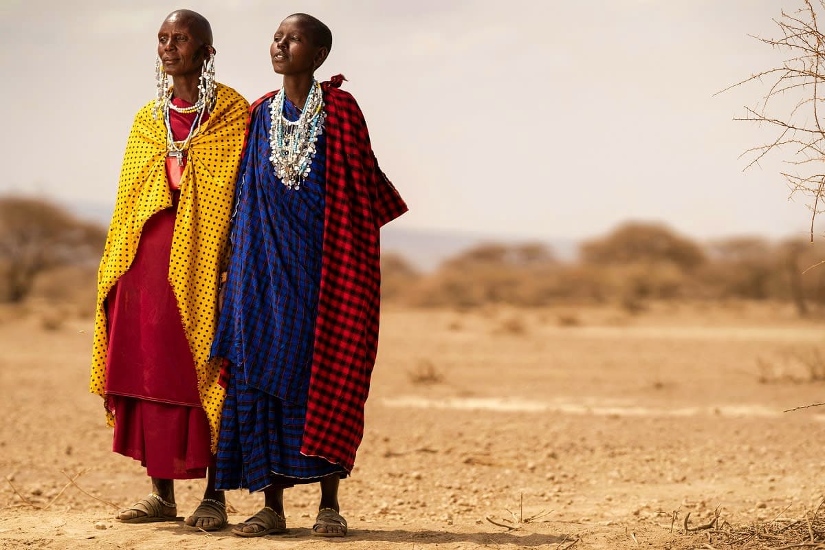 Two Maasai women in traditional colorful clothing standing in an arid savanna landscape in Kenya.