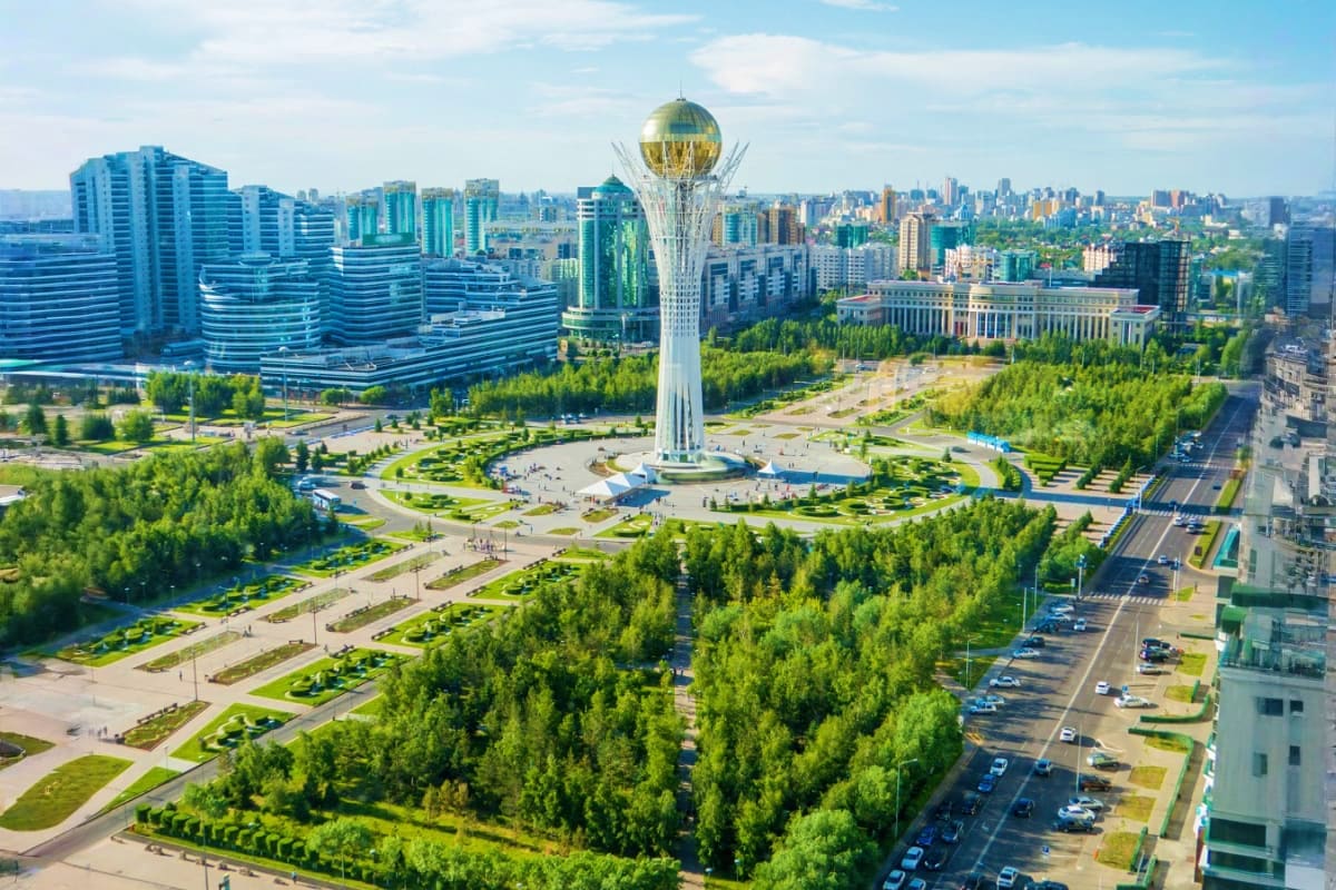 Aerial view of Bayterek Tower with golden sphere in Astana, surrounded by modern blue buildings, green parks, and tree-lined 