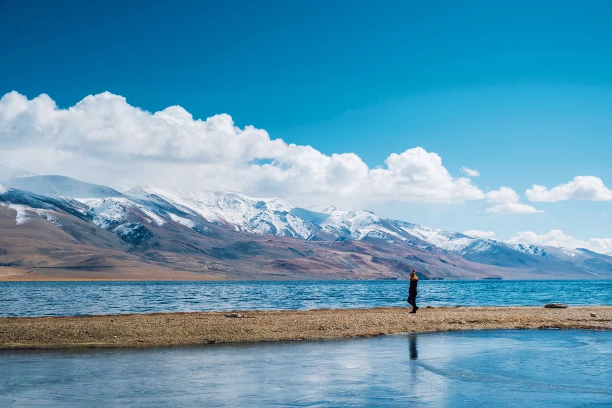 Person standing on beach beside turquoise alpine lake with snow-capped Himalayan mountains under blue sky with white clouds