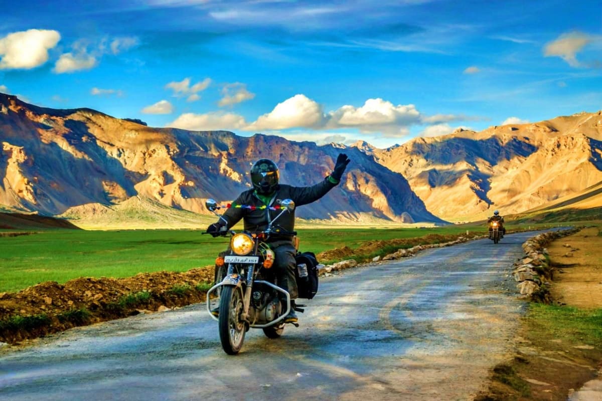 Motorcyclist with raised arm riding on mountain road through Ladakh valley with snow-capped peaks and green fields in backgro