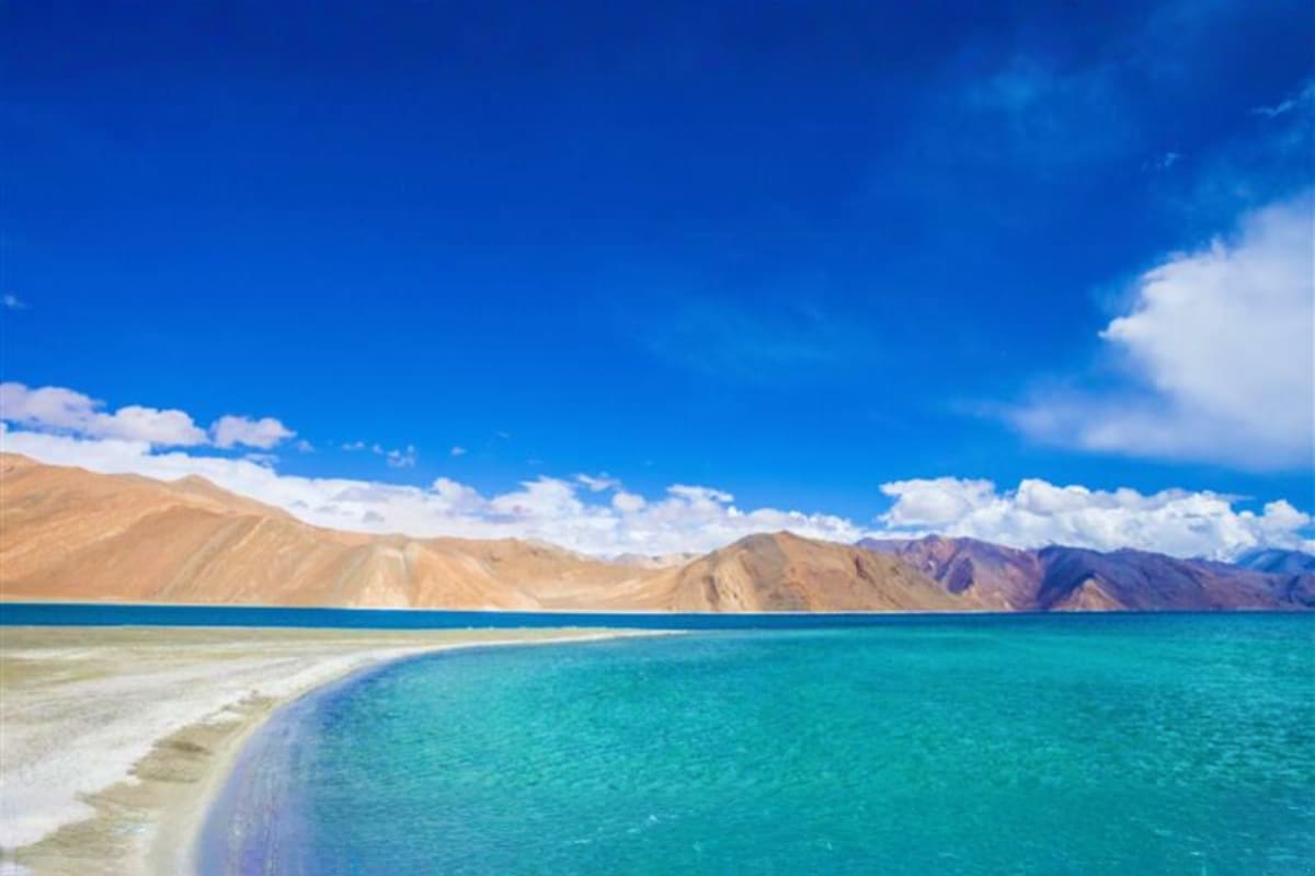 Turquoise waters of Pangong Lake with white sandy beach and barren mountain ranges under blue sky with white clouds in Ladakh