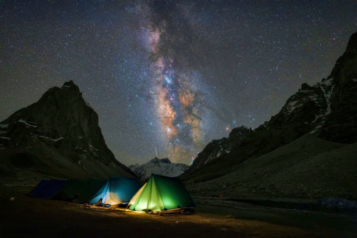 Illuminated tents pitched in mountain valley beneath Milky Way galaxy and snow-capped peaks at night during Ladakh camping ex