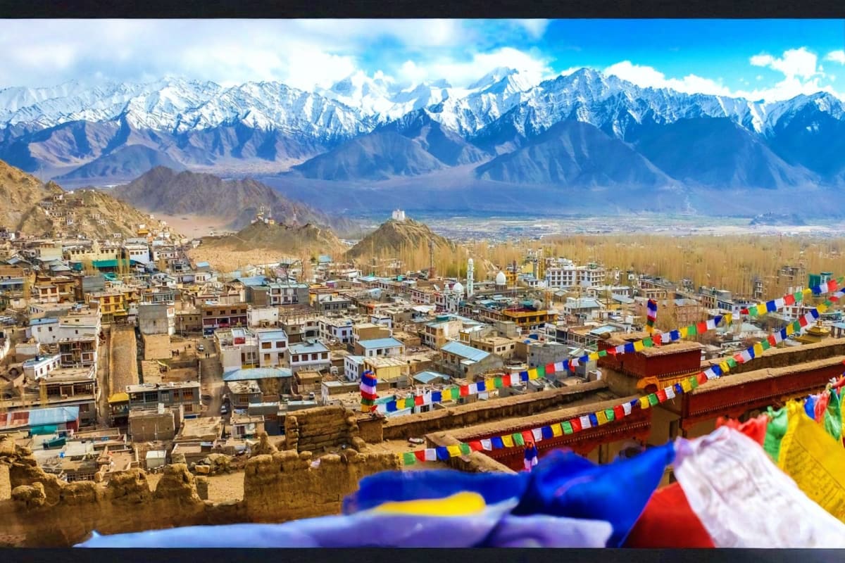 Leh city with snow-capped Himalayan mountains, colorful Buddhist prayer flags in foreground, and traditional Ladakhi architec