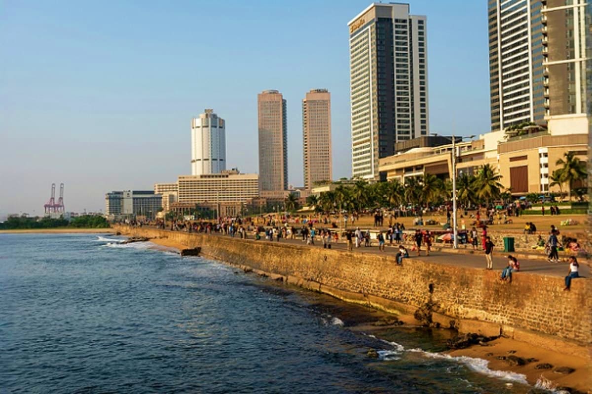 Crowded beachfront promenade with stone seawall, sandy shore, and high-rise buildings along the waterfront under clear sky