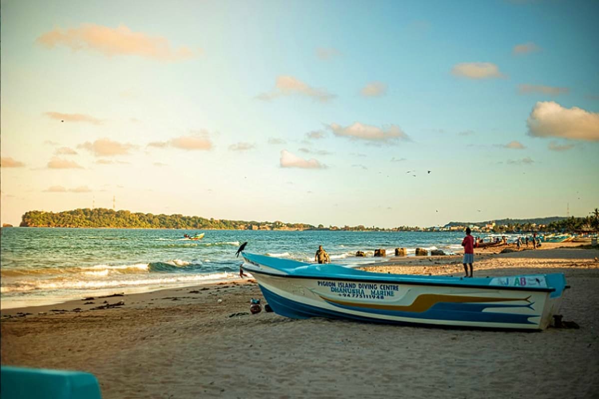 Blue and white fishing boat on sandy beach with swimmers and forested island in background, Mirissa, Sri Lanka
