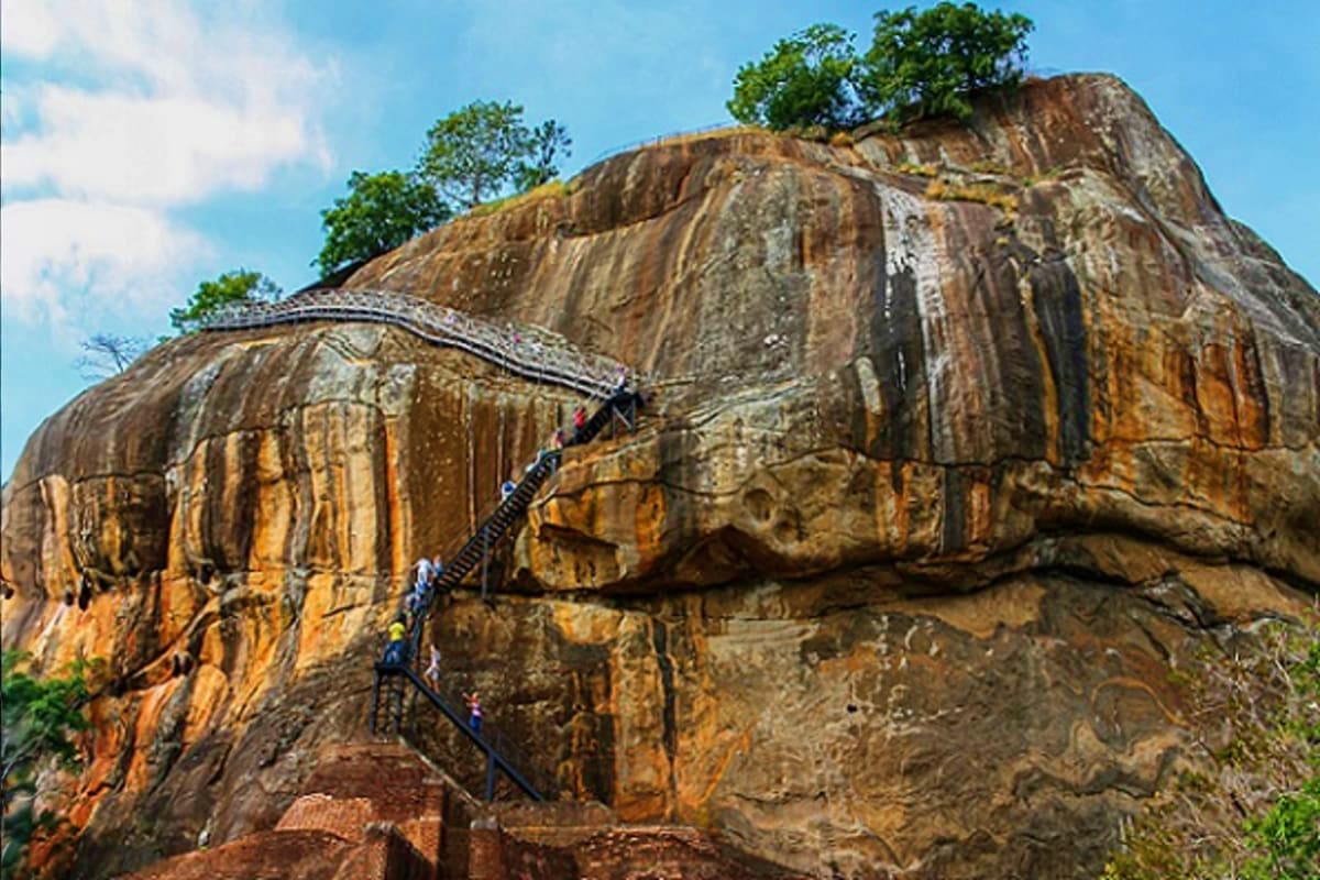 Tourists climbing metal staircase on large rocky outcrop with ancient rock formations and green vegetation against blue sky