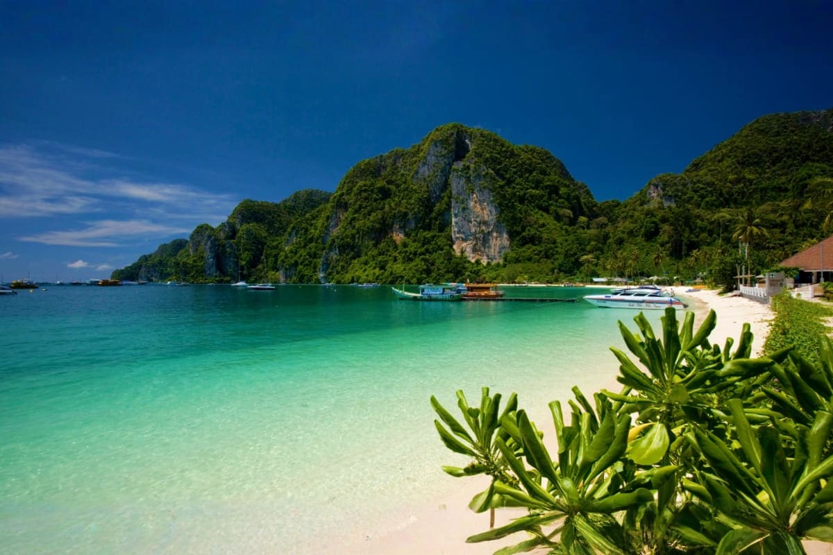 Tropical beach in Thailand with turquoise waters, white sand, limestone cliffs, and traditional longtail boats anchored in th