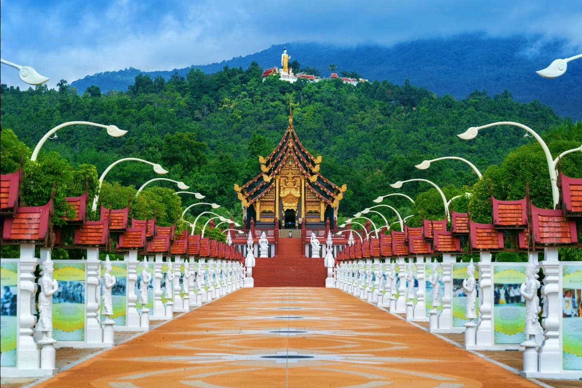 Symmetrical wooden bridge with white railings and decorative arches leading to ornate golden Thai temple with red roofs, surr